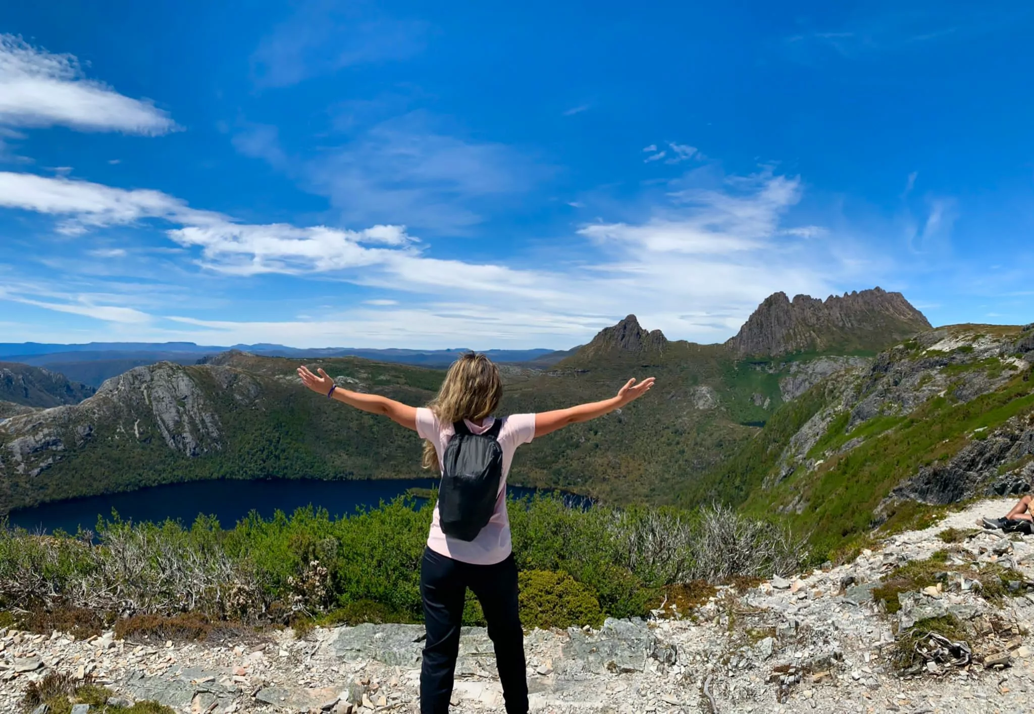A girl with arms stretched wide sstands on Marion's Lookout with Cradle Mountain and Dove Lake in the distance behind her. She wears a tshirt and backpack and the sky is blue.