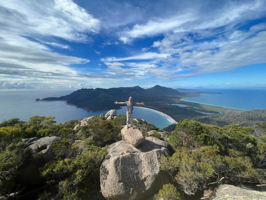 A man with arms stretched wide stands on a boulder on Mount Amos with views of Wineglass Bay and Freycinet National Park stretch into the distance.