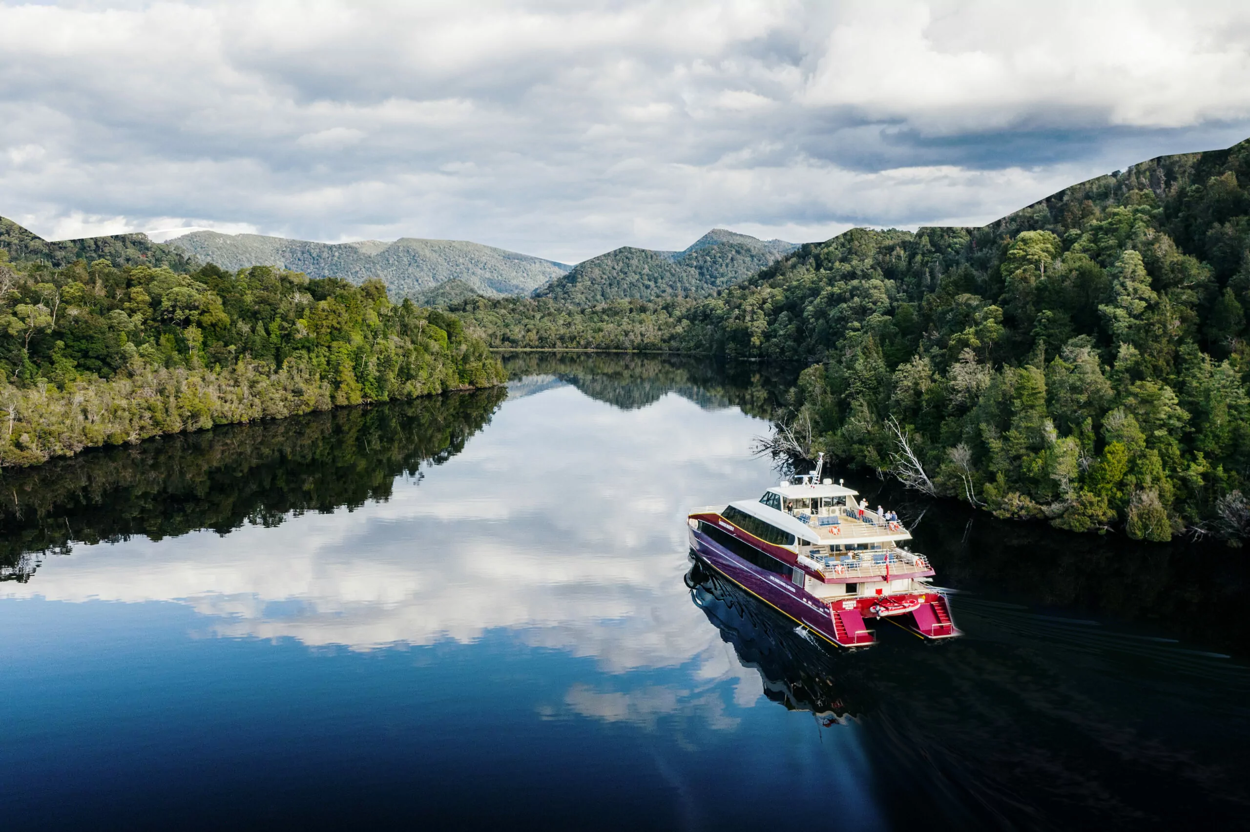 A red boat glides up the gordon river surrounded by rainforest. The sky is reflected in the calm waters.