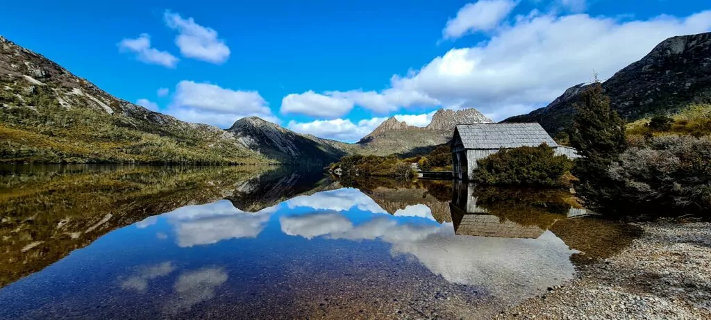 The sky reflects in Dove Lake with the boatshed in the foreground and Cradle Mountain clear in the background.