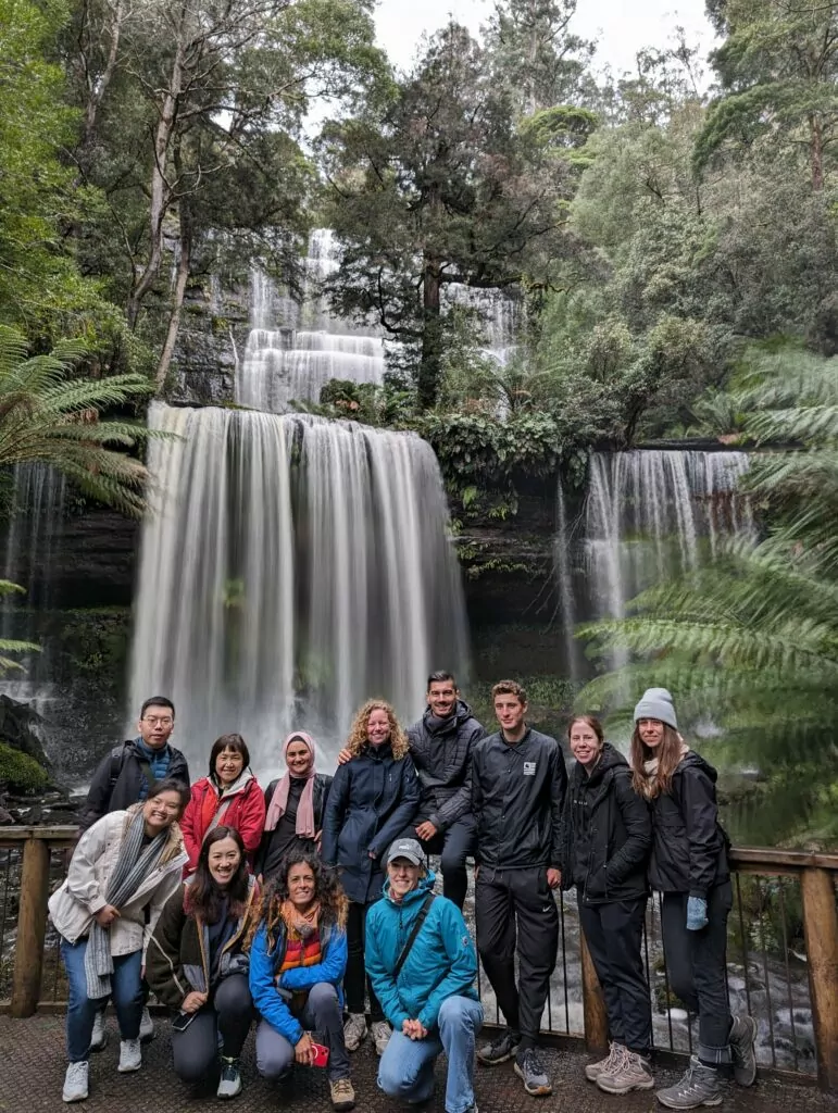 A group poses at Russell Falls surrounded by forest. The falls cascade beautifully behind them