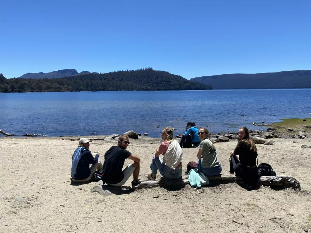 Small group of young people sit on the sands of Lake St Clair enjoying sunshine and lunch